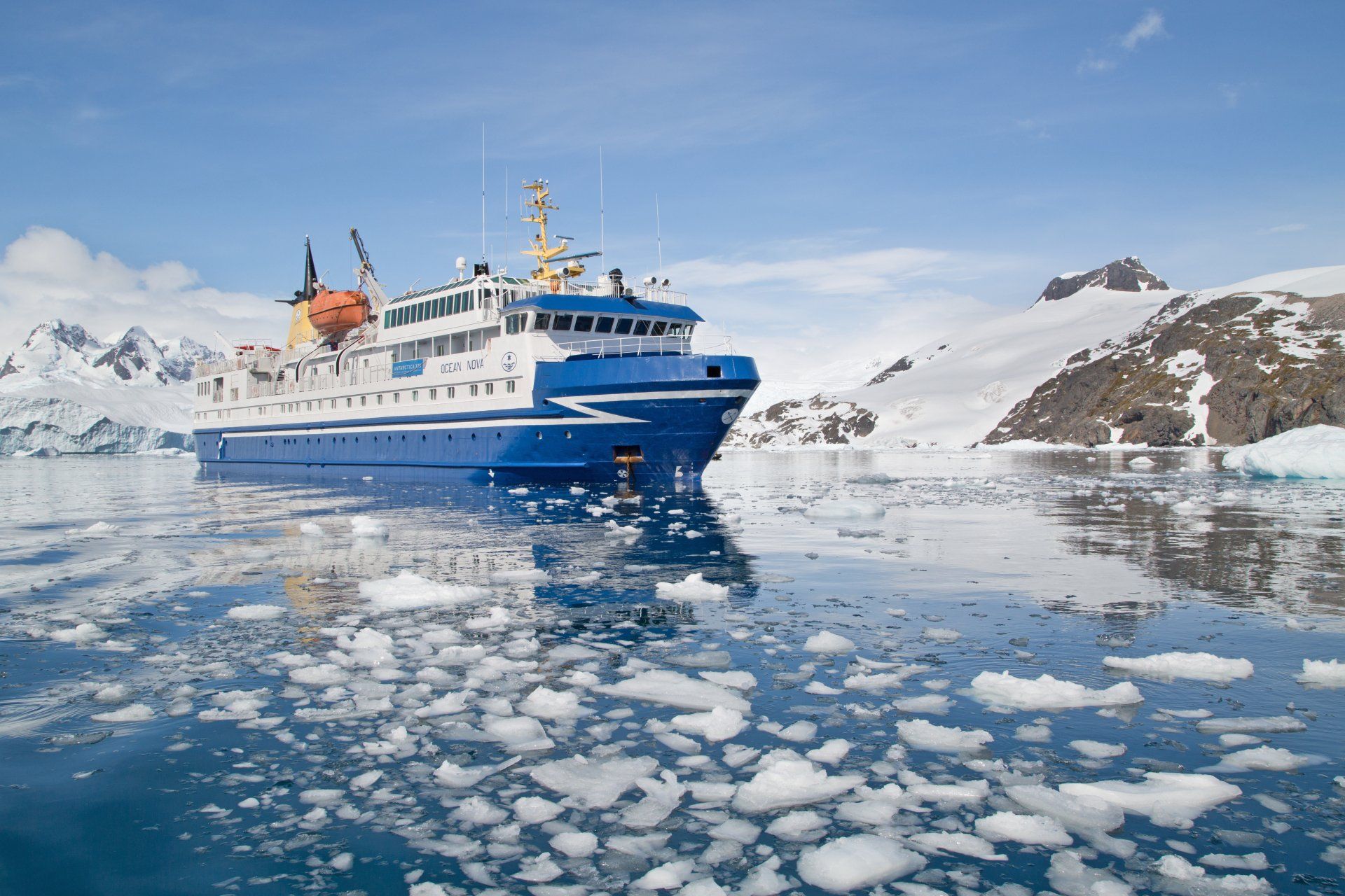 Blue and white ship sailing in icy water surrounded by icebergs and snow-covered mountains under a clear blue sky.