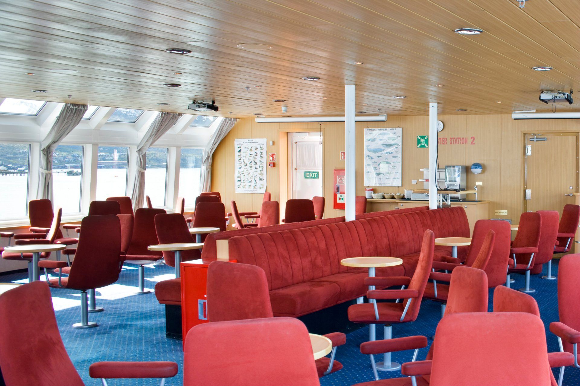 Interior of a ferry with red seating, tables, and a curved red sofa on blue carpet, under a light wood ceiling.