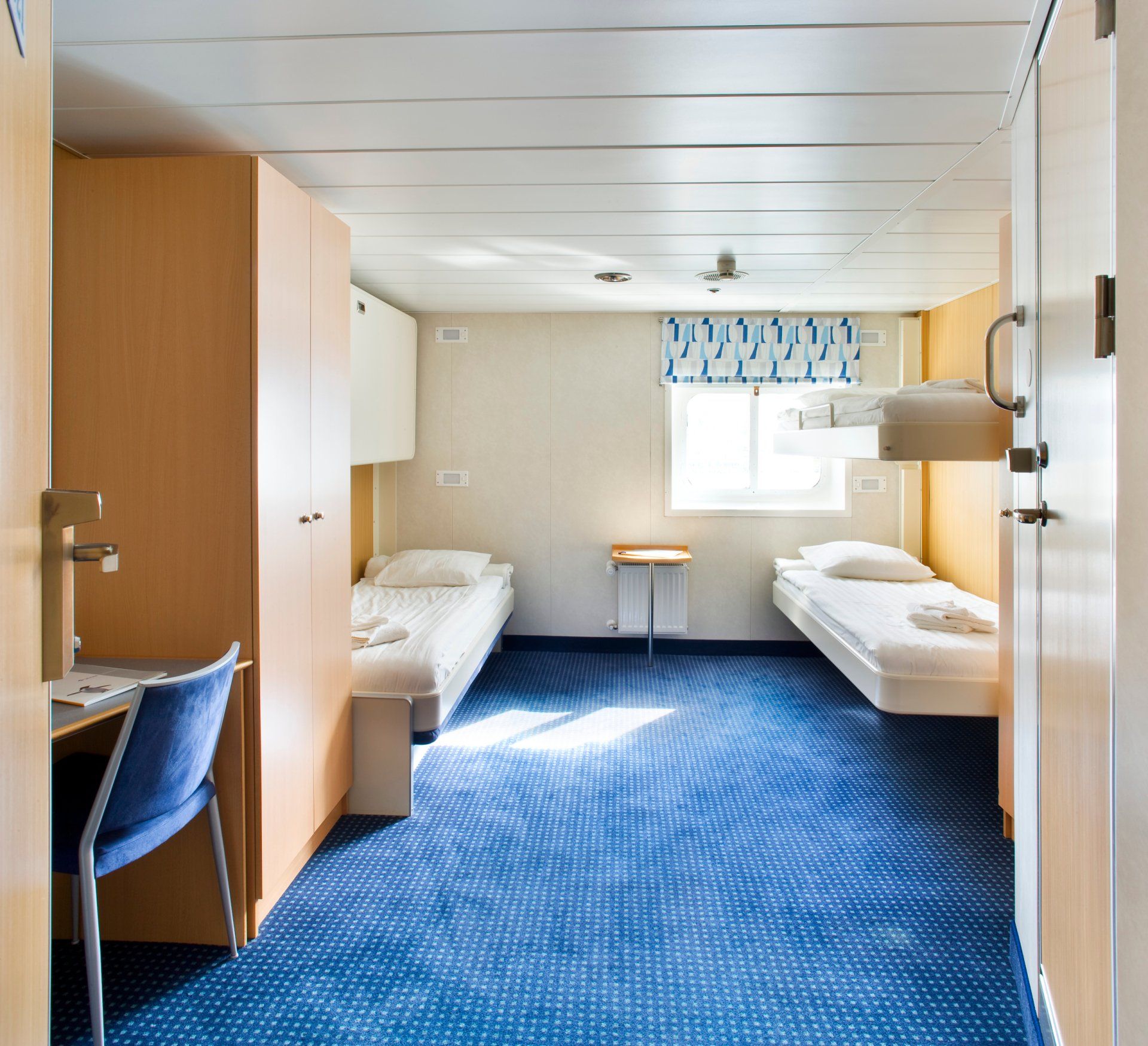 Cabin interior with blue carpet, two beds, a desk, and a window with a blue-patterned shade.