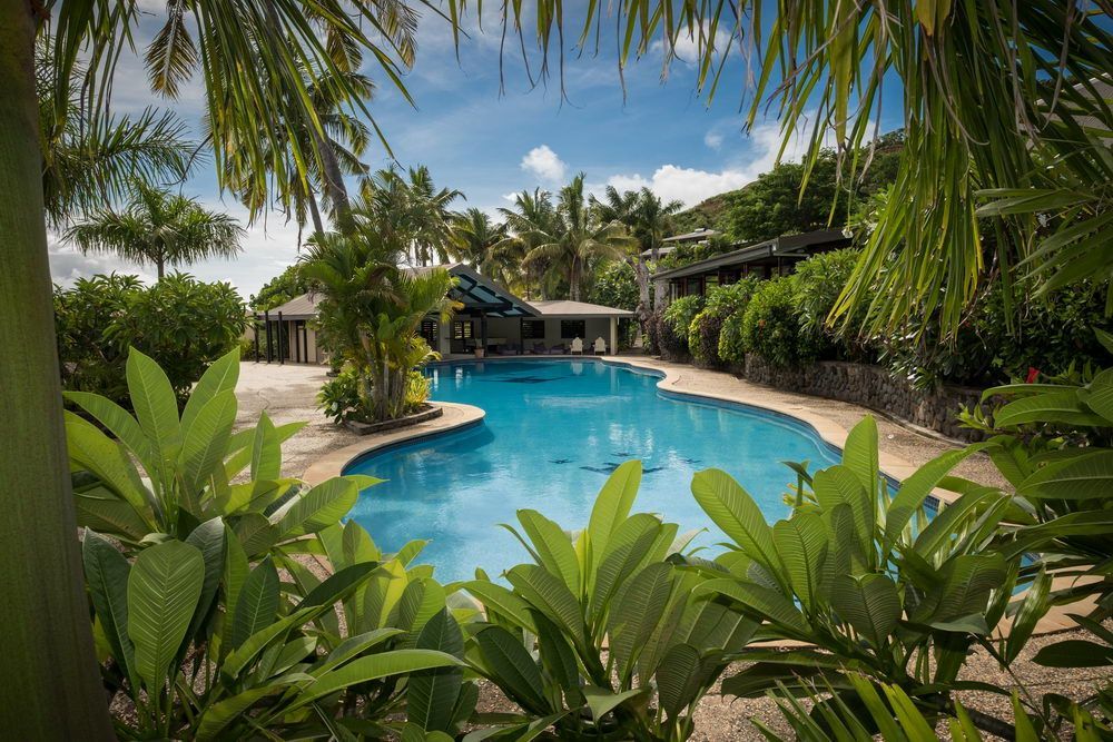 Swimming pool surrounded by tropical plants and buildings, under a blue sky.