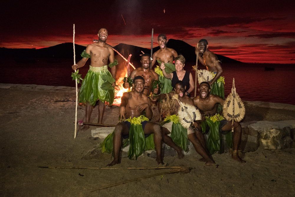 Group of people, including one woman, pose by fire on beach at sunset, wearing leaf skirts and holding spears.