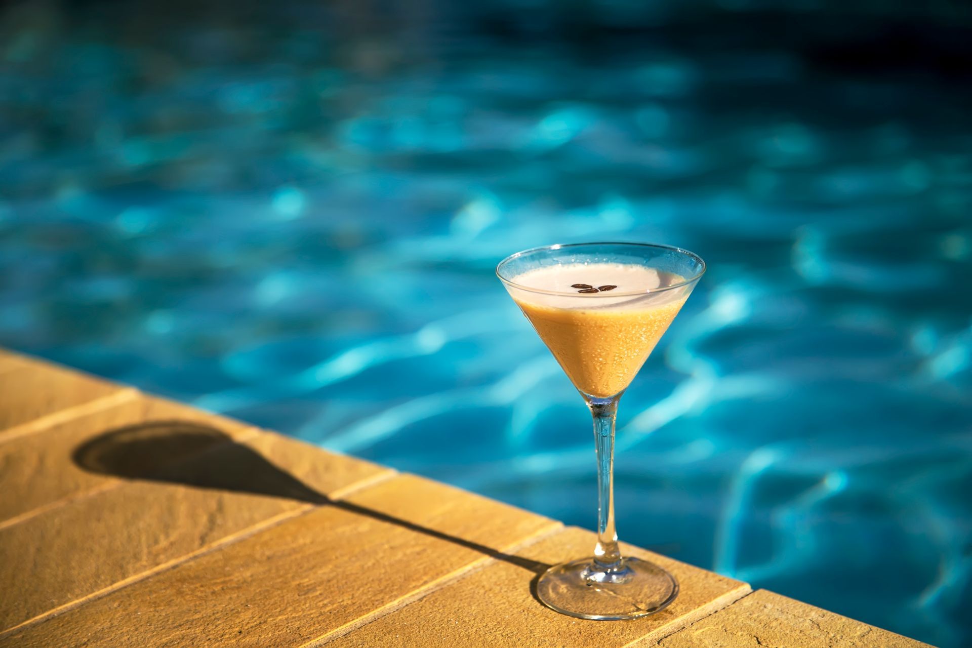 Cocktail in a martini glass on a pool deck, with the blue water of the pool in the background.