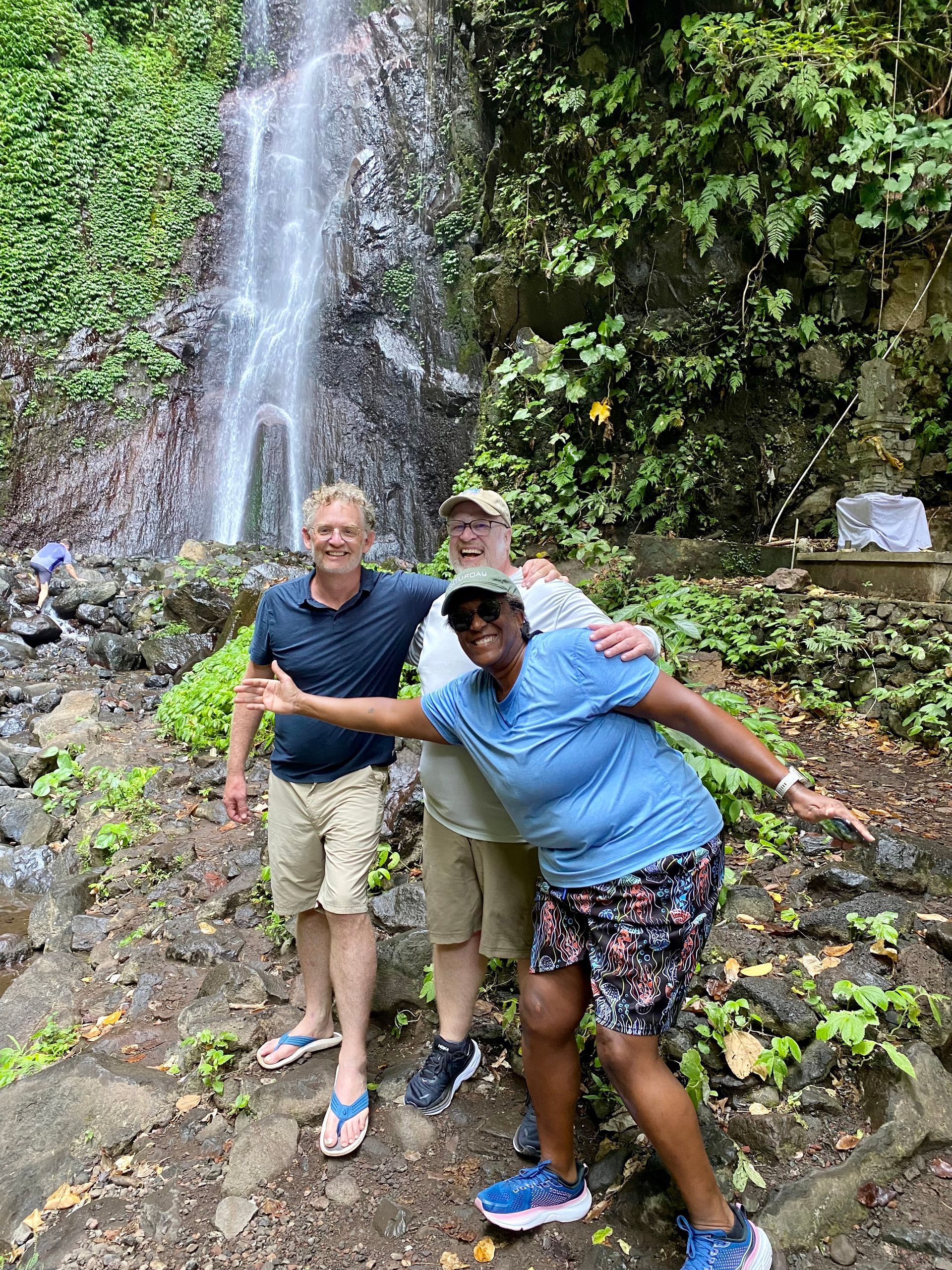 Three people posing by a waterfall. One person gestures with arms outstretched. Lush greenery surrounds them.