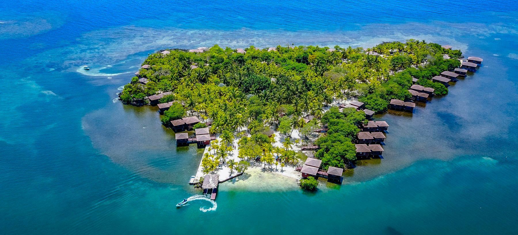 Aerial view of a tropical island with overwater bungalows in turquoise water. Lush green trees cover the island.
