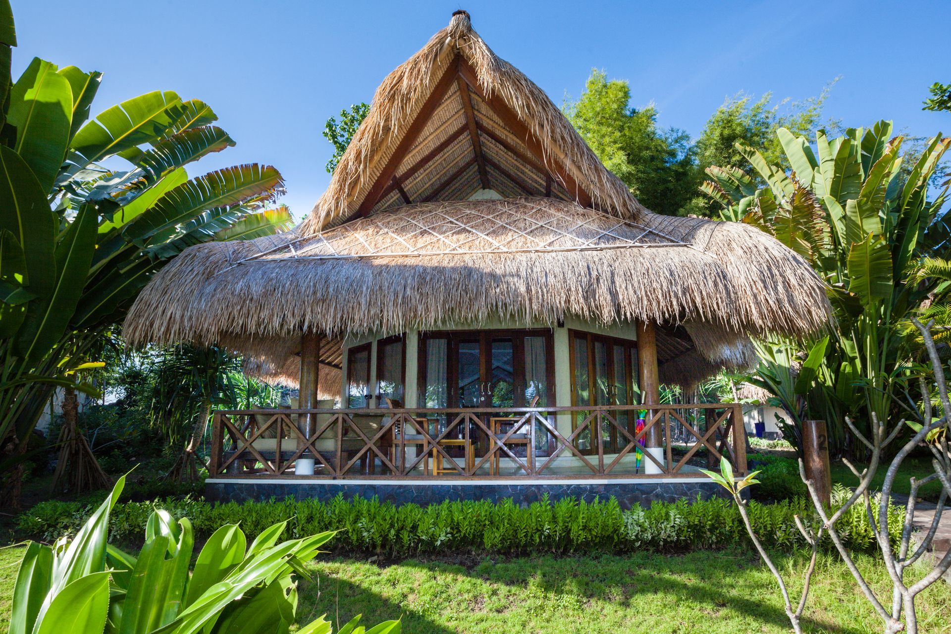 Thatched-roof bungalow with wooden balcony, surrounded by lush green plants under a clear blue sky.