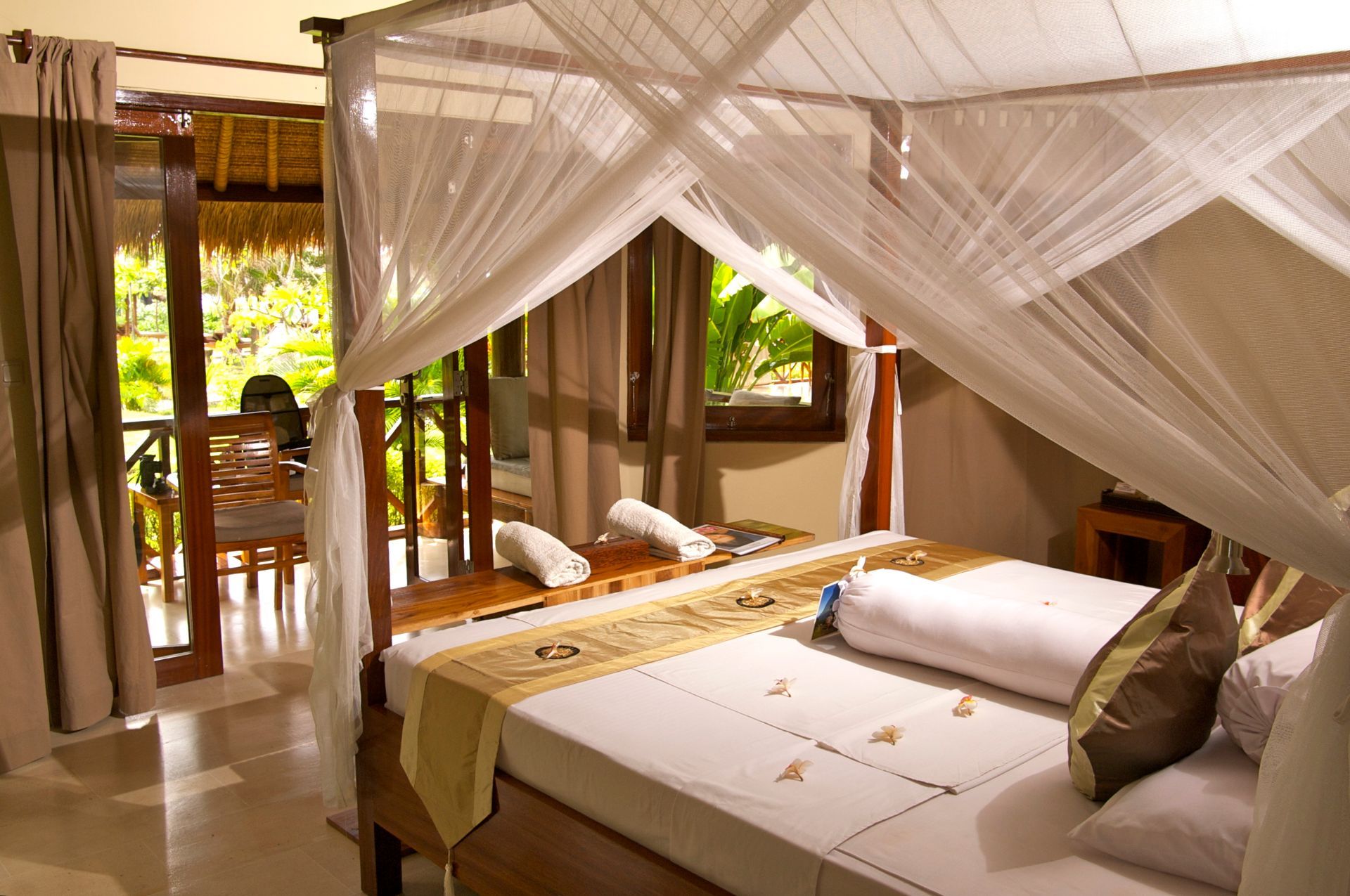 Bedroom with canopy bed, open to a balcony with greenery. White linens, tan accents.
