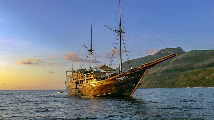 Wooden sailing ship on calm water with mountain backdrop at sunset.