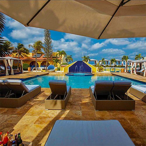 Pool with lounge chairs under a large umbrella, with a building and palm trees in the background.