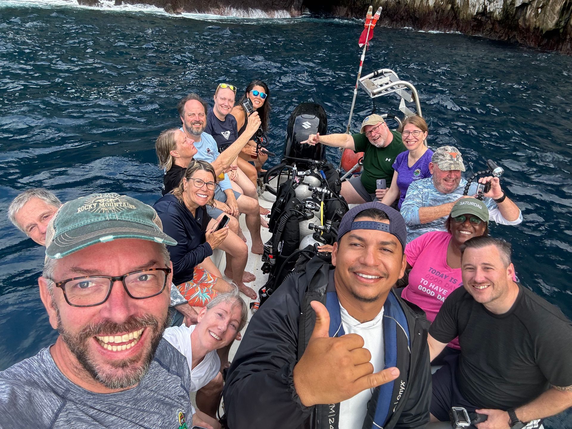 Group of people on a boat, smiling for a photo; blue water and rocky shore in the background.