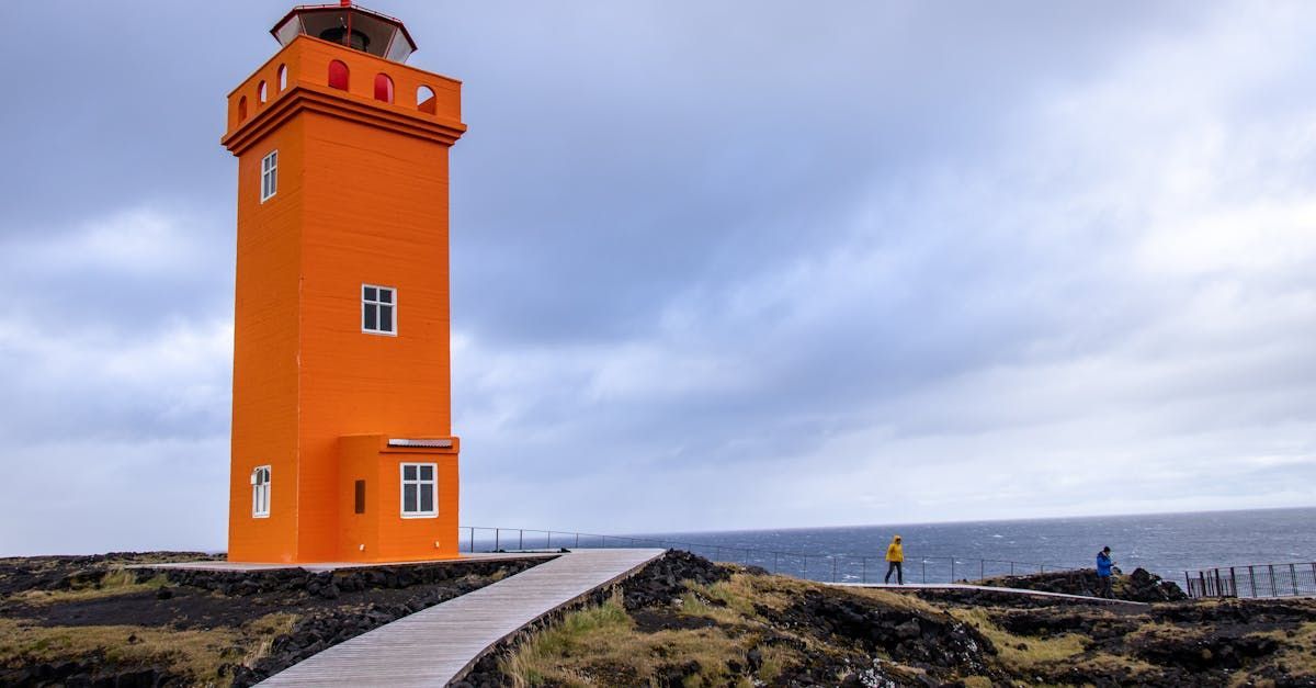 Orange lighthouse on a rocky coast with a walkway. Cloudy sky, ocean, and a few people in the distance.