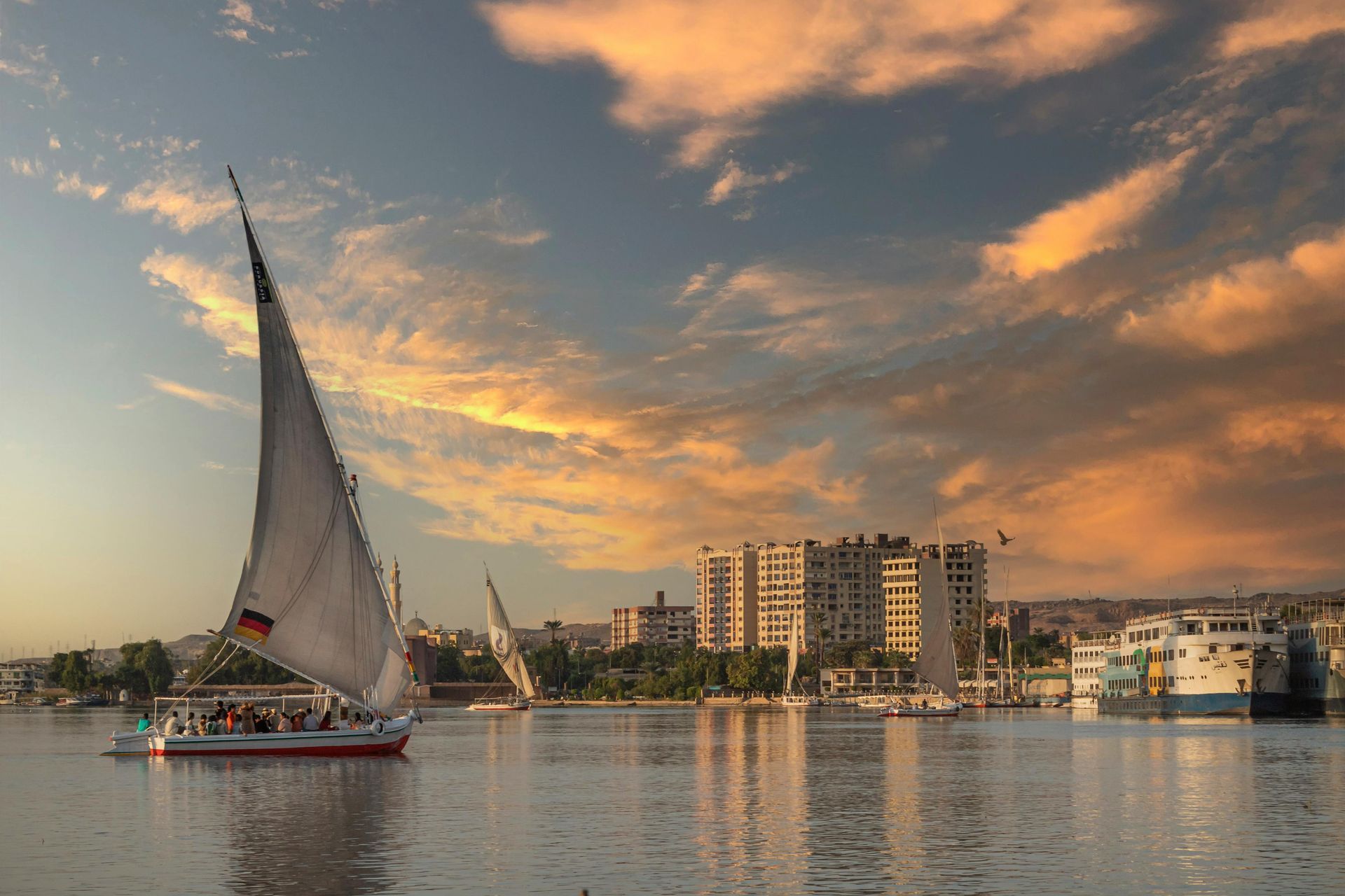 Boats sail on a river reflecting sunset colors; buildings line the shore under a partly cloudy sky.
