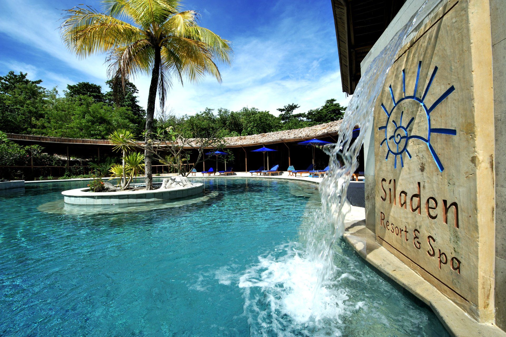 Swimming pool at Siladen Resort & Spa with water feature and resort signage. Lush greenery and blue sky.