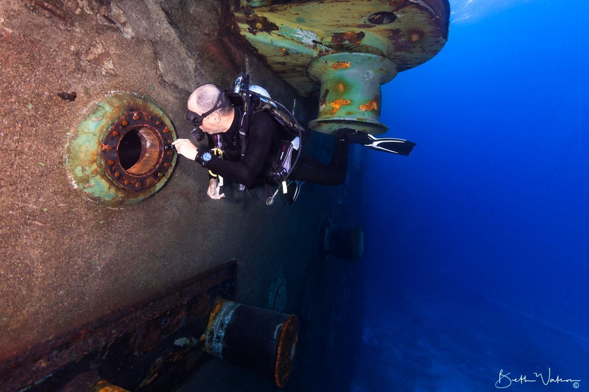 Scuba diver examines rusted, cylindrical object on the side of a sunken vessel in deep blue water.