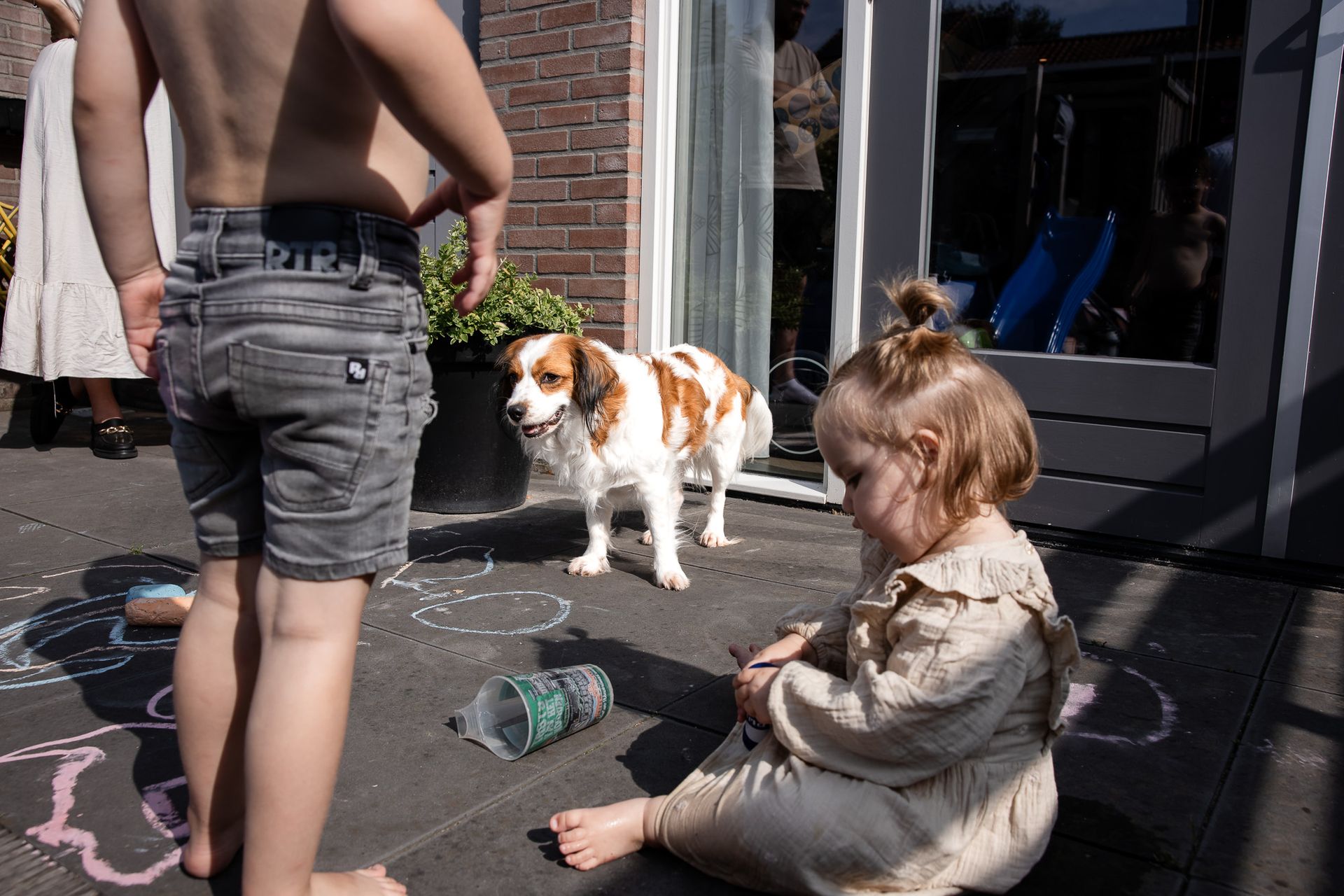 Twee jonge kinderen spelen buiten met stoepkrijt op het terras, met de hond erbij