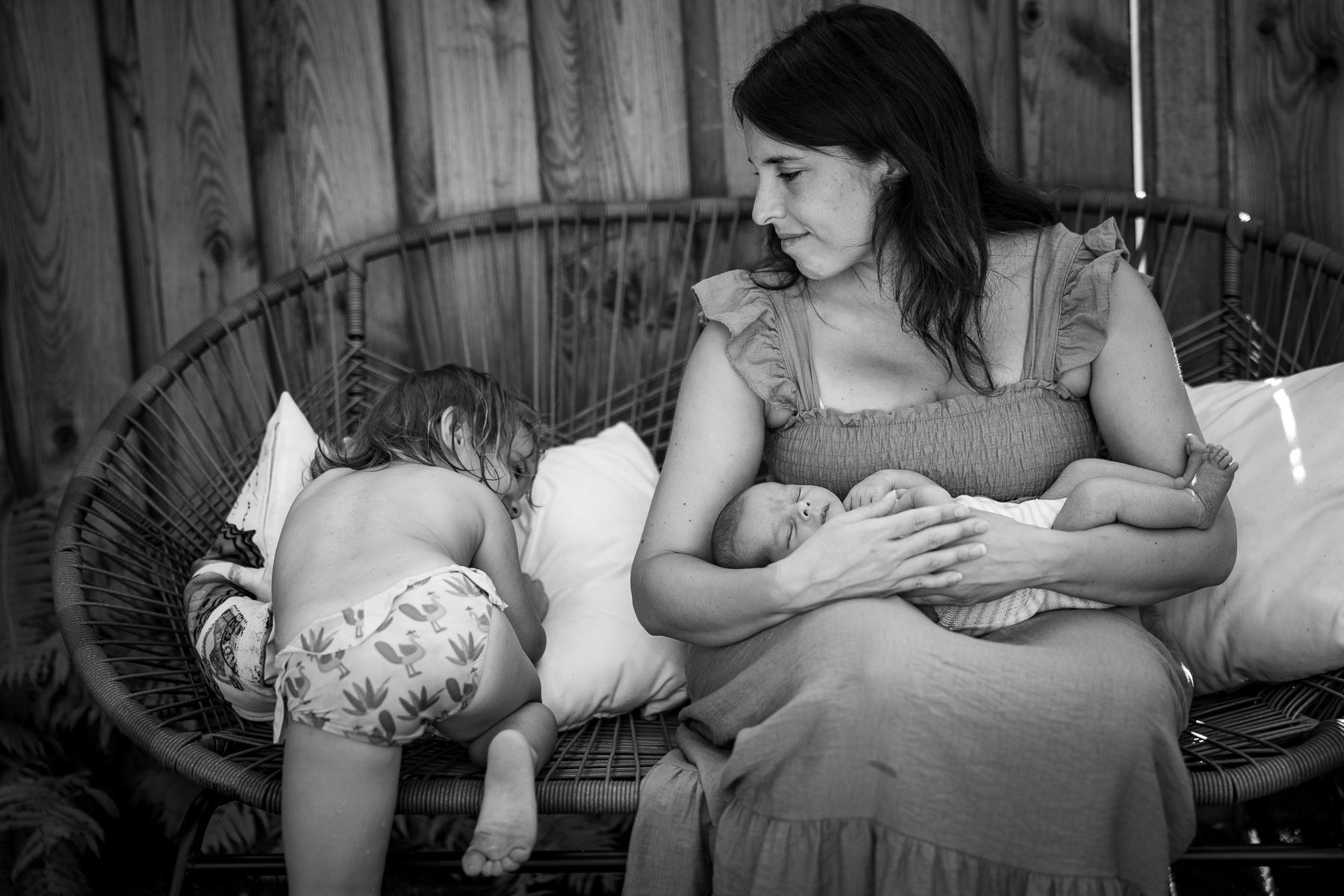 Moeder zit met haar pasgeboren baby terwijl een peuter naast haar op de bank ligt.