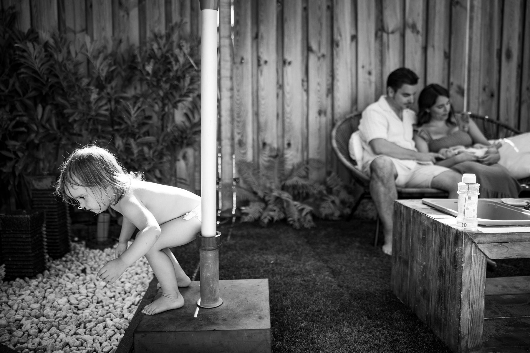 Peuter speelt in de tuin terwijl ouders met hun baby samen zitten op de achtergrond.