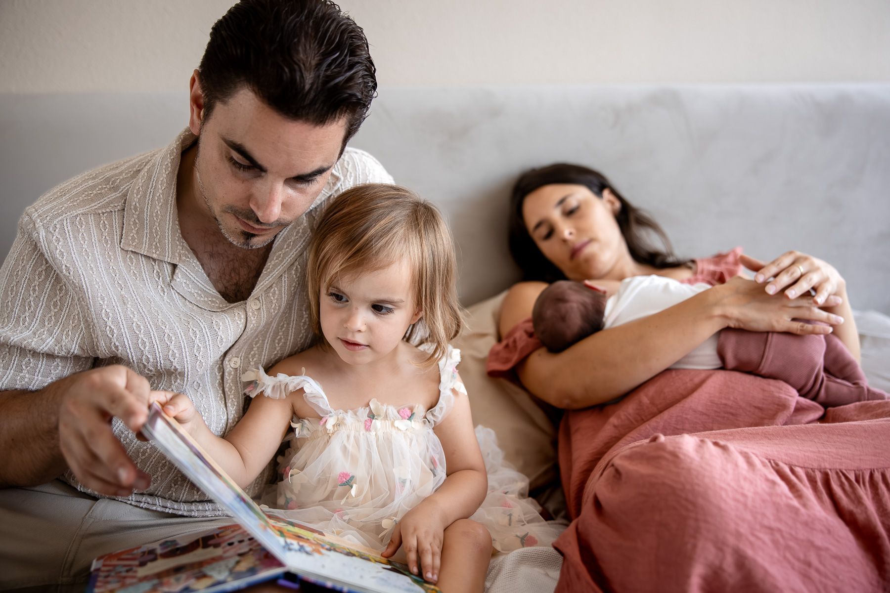 Vader leest voor aan peuter terwijl moeder met pasgeboren baby op bed ligt tijdens een Day in the Life reportage