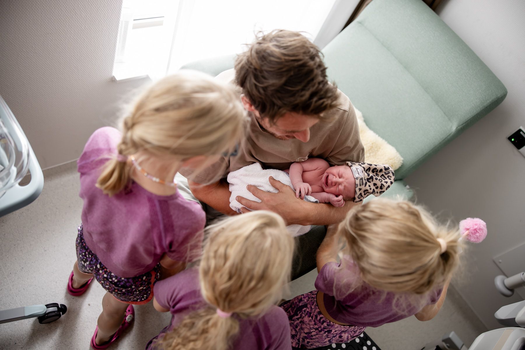 Vader houdt zijn pasgeboren baby vast terwijl zusjes nieuwsgierig komen kijken.