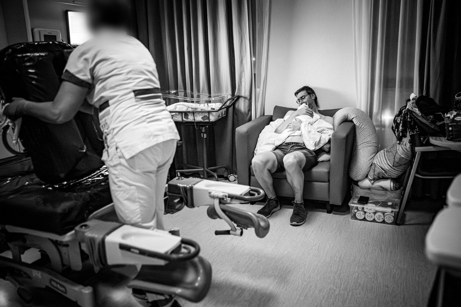 Vader zit met zijn pasgeboren baby in een stoel in de ziekenhuiskamer terwijl een verpleegkundige de kamer schoonmaakt.