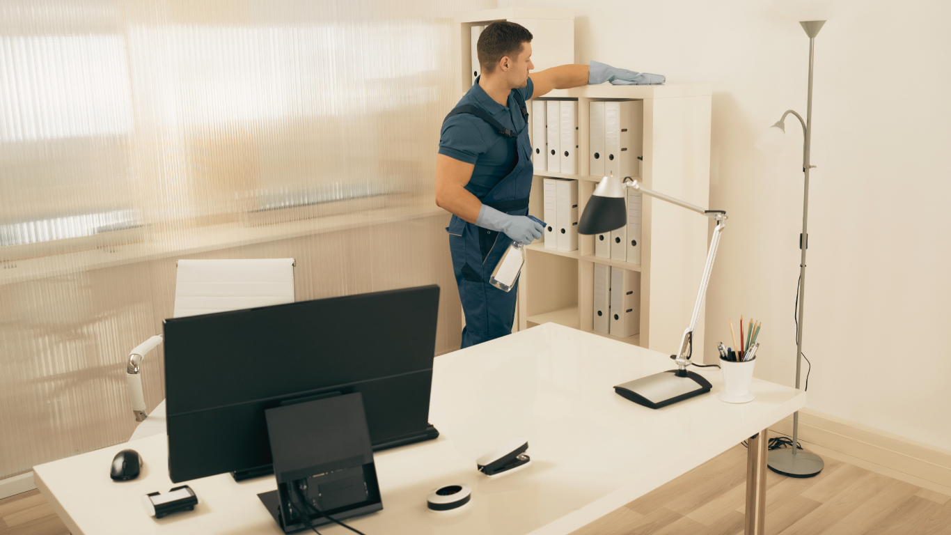Person in blue scrubs and gloves cleaning an office, wiping a shelf with a spray bottle.