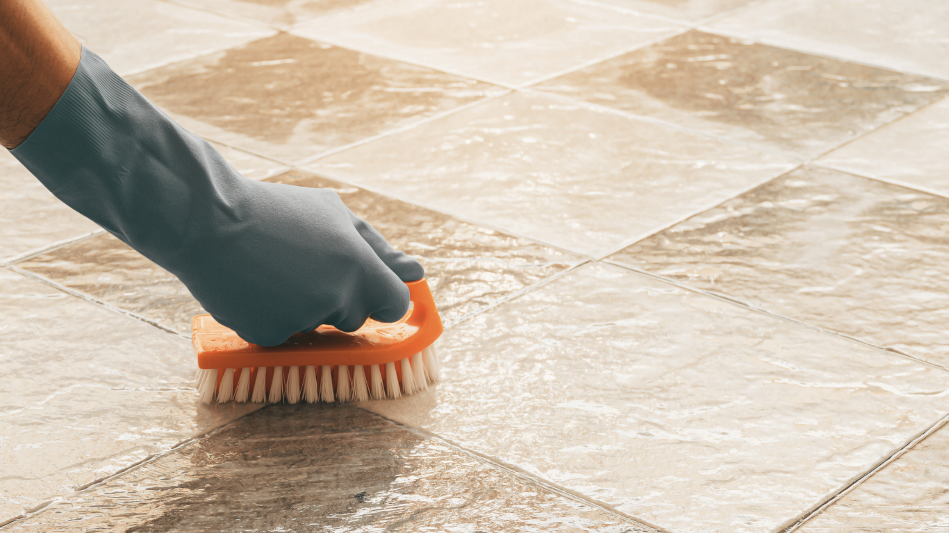Gloved hand scrubbing tiled floor with an orange brush.