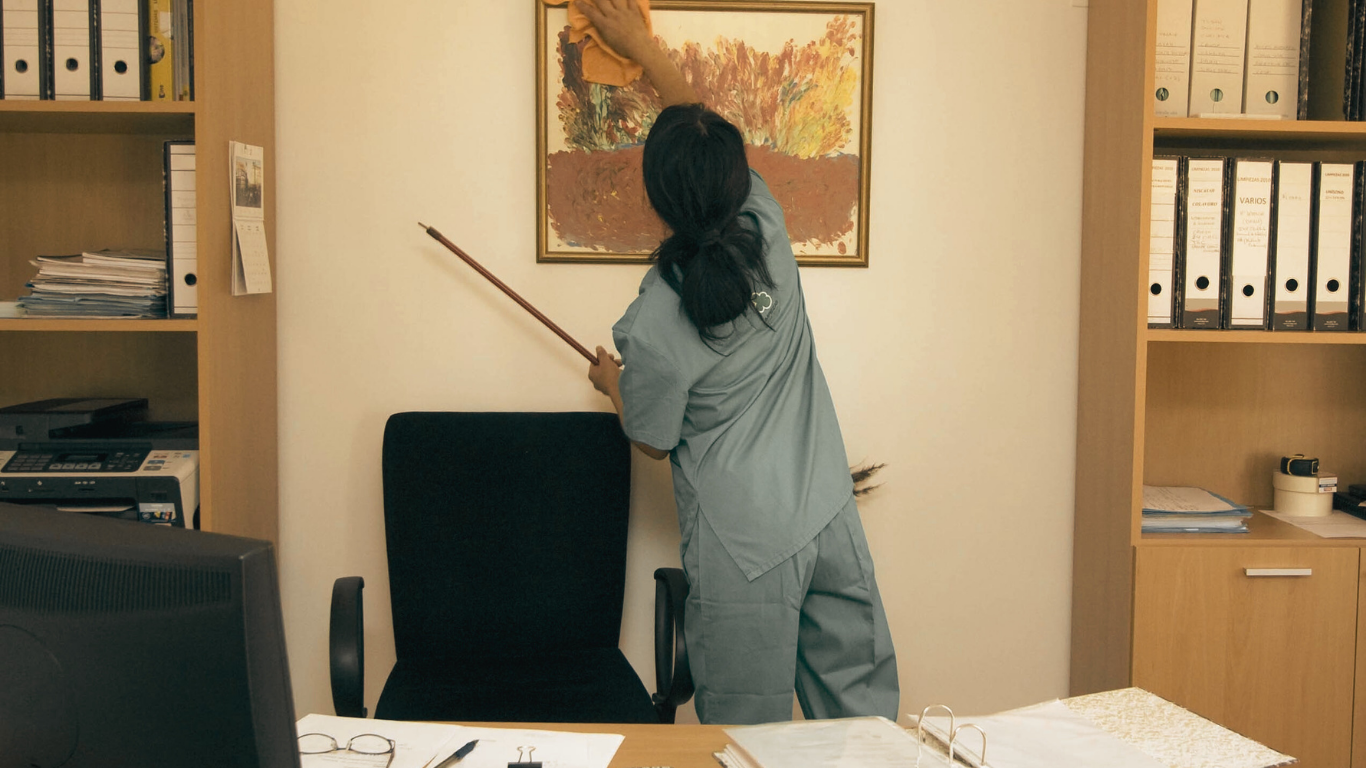 Person in blue scrubs uses a stick to adjust a painting in an office.