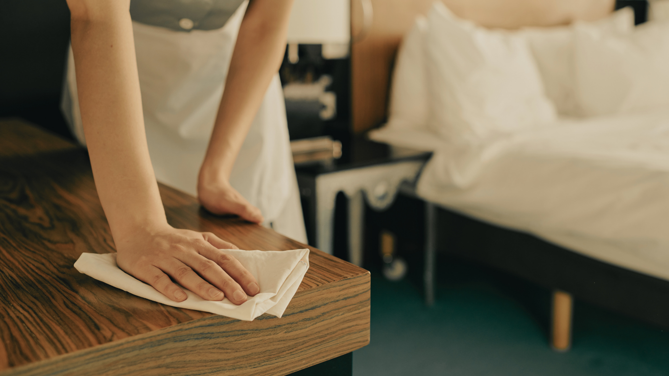 Maid wiping down a wooden table in a hotel room with a white cloth. Bedroom in background.