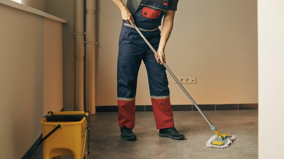 Person mopping a gray floor in a room. They wear blue overalls with red accents and black shoes; a yellow bucket sits nearby.