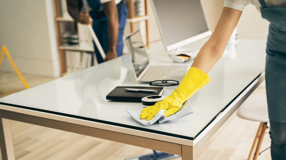 Person in yellow glove wiping a clean, white desk. Another person cleans in the background. Office setting.