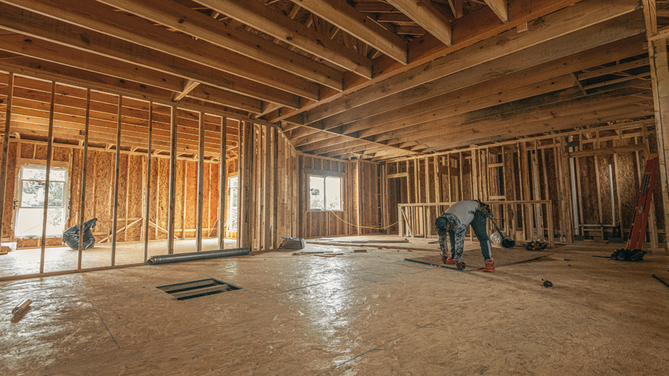 Interior view of a building under construction, with exposed wooden framework. A person works on the floor.