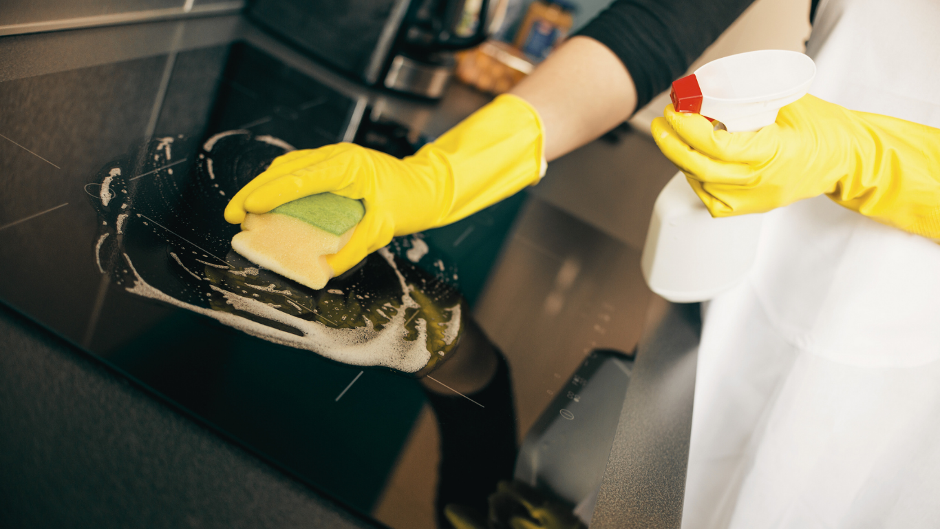 Person wearing yellow gloves cleans a black stovetop with a sponge and spray bottle.
