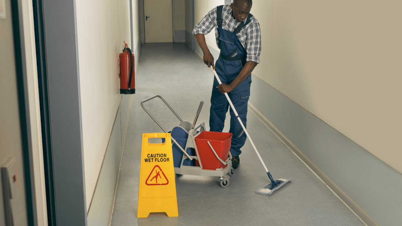 Man mopping a hallway floor, caution sign and cleaning cart nearby.