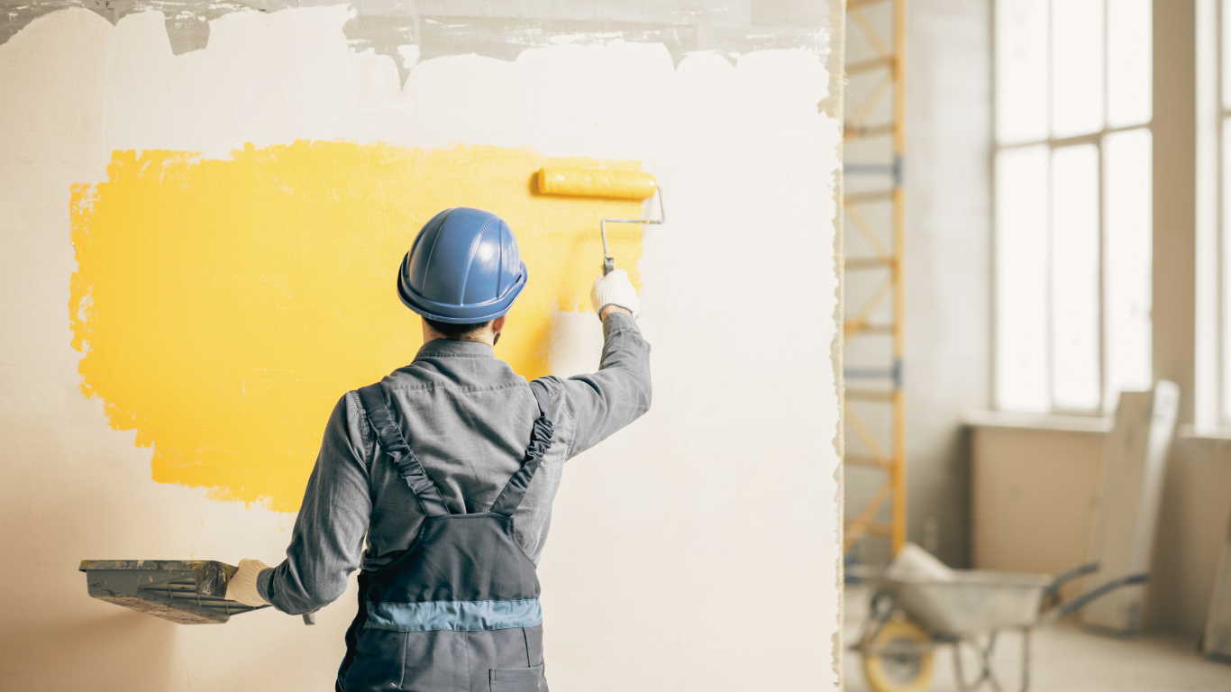 Person in blue hard hat painting a yellow rectangle on a white wall with a paint roller.
