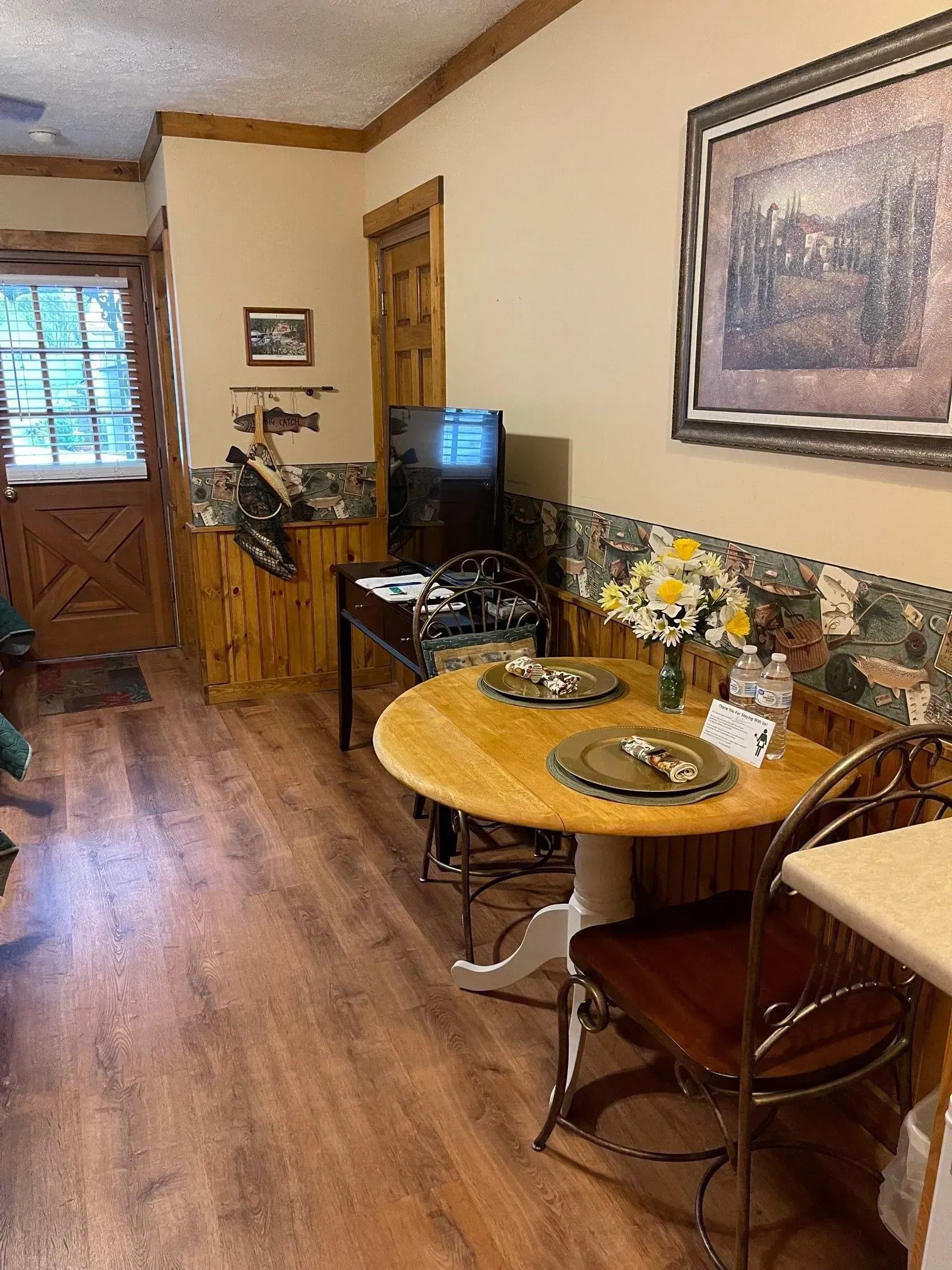 Cozy dining area: round table, two chairs, wooden floor, TV, art, door to the outside.