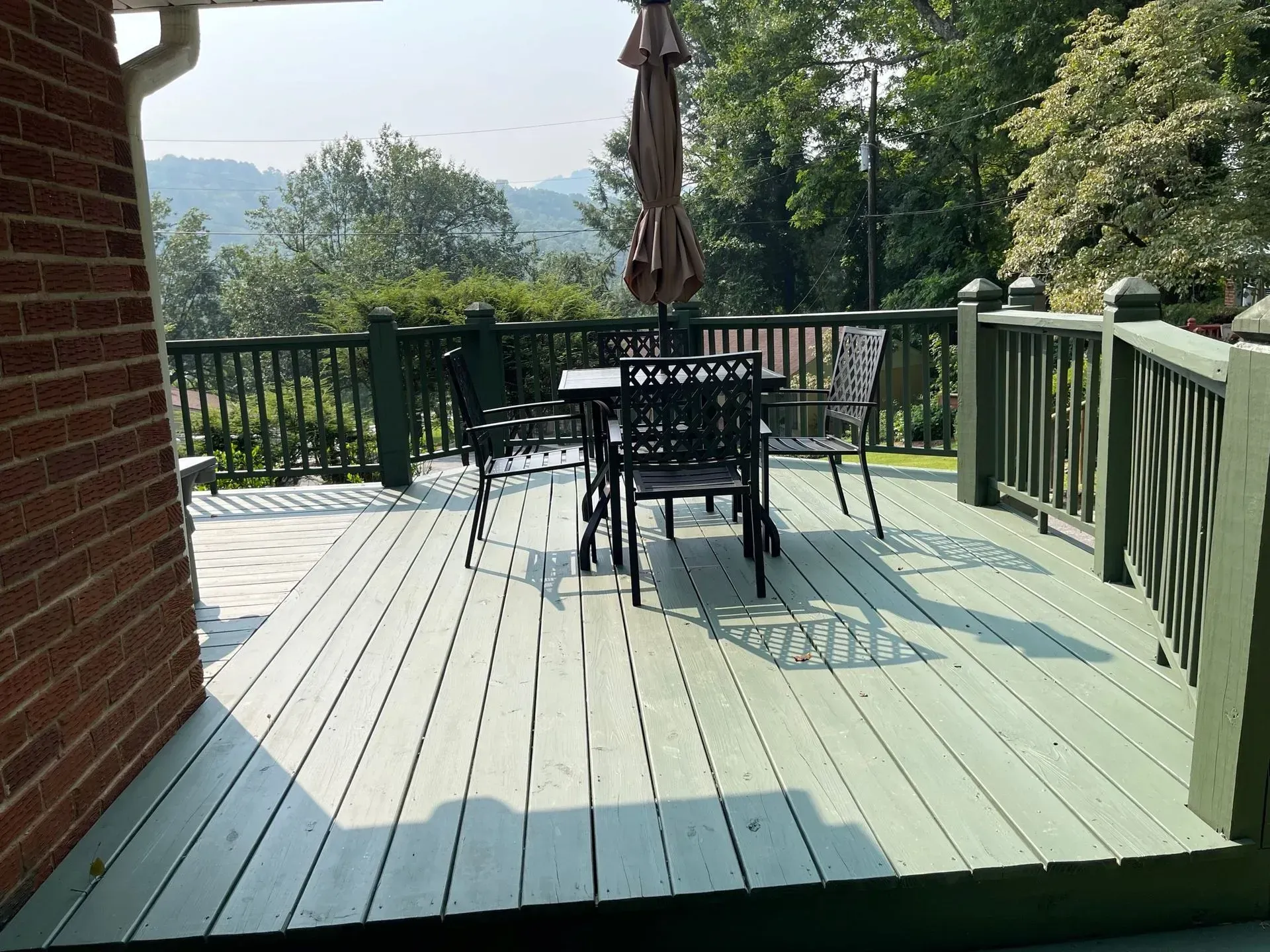 Green deck with dark metal table, chairs, umbrella, and a brick wall with a scenic, tree-filled background.
