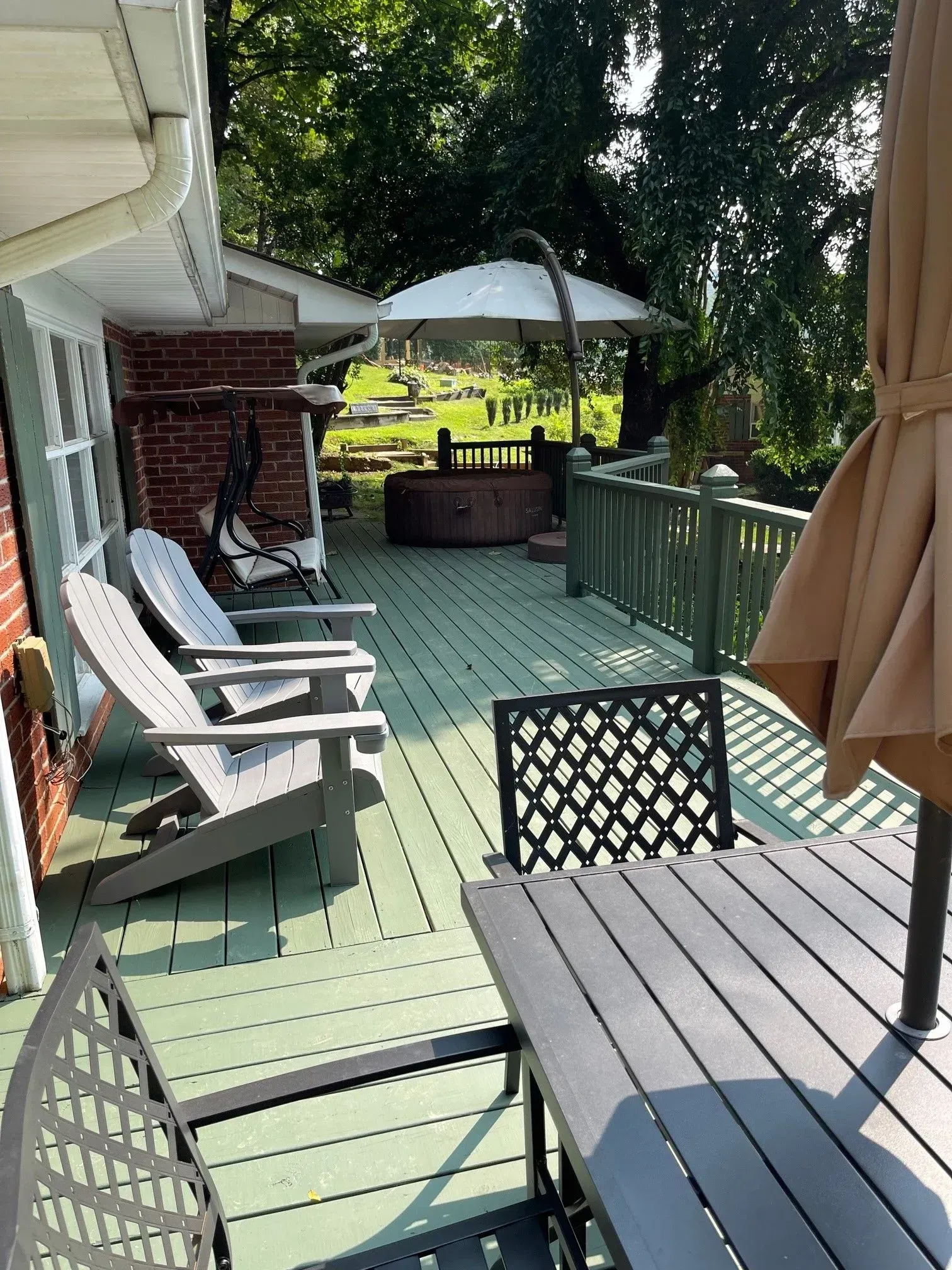 Deck with Adirondack chairs, table, and umbrella, leading to a hot tub and gazebo in a sunny backyard.