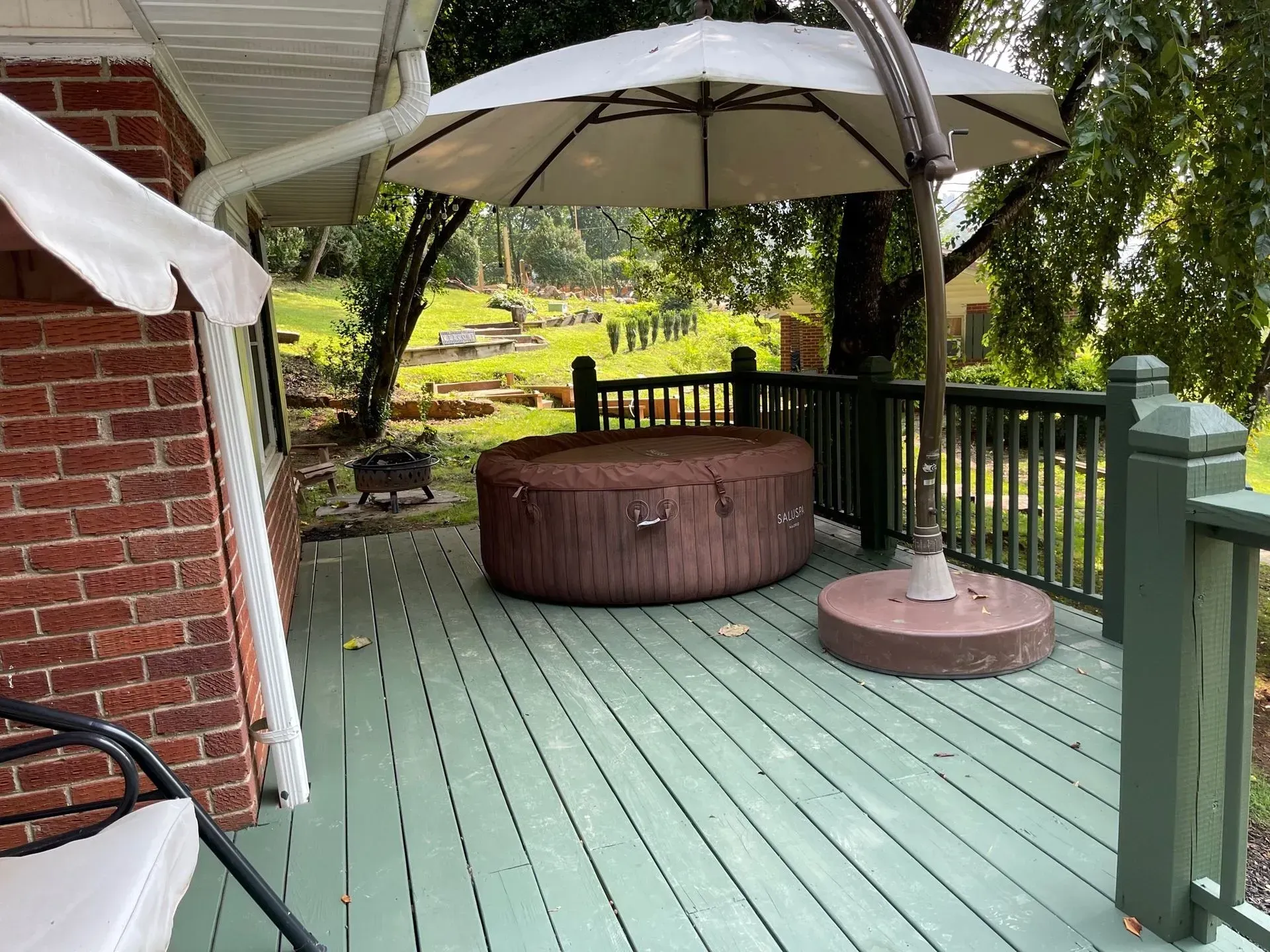 A hot tub on a wooden deck, shaded by an umbrella, next to a brick house.