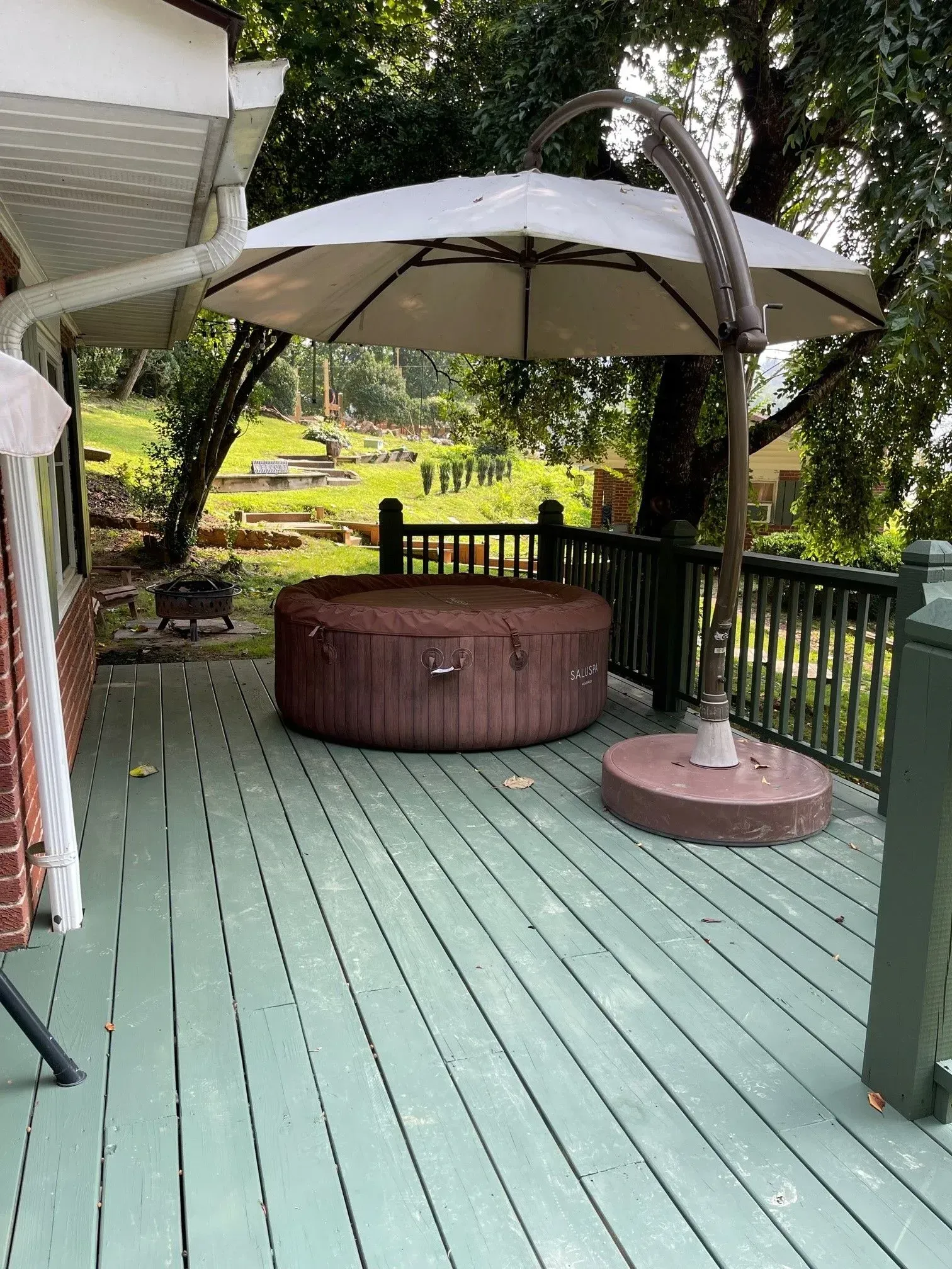 Wooden deck with brown hot tub, offset umbrella, and green railing. Trees in background.