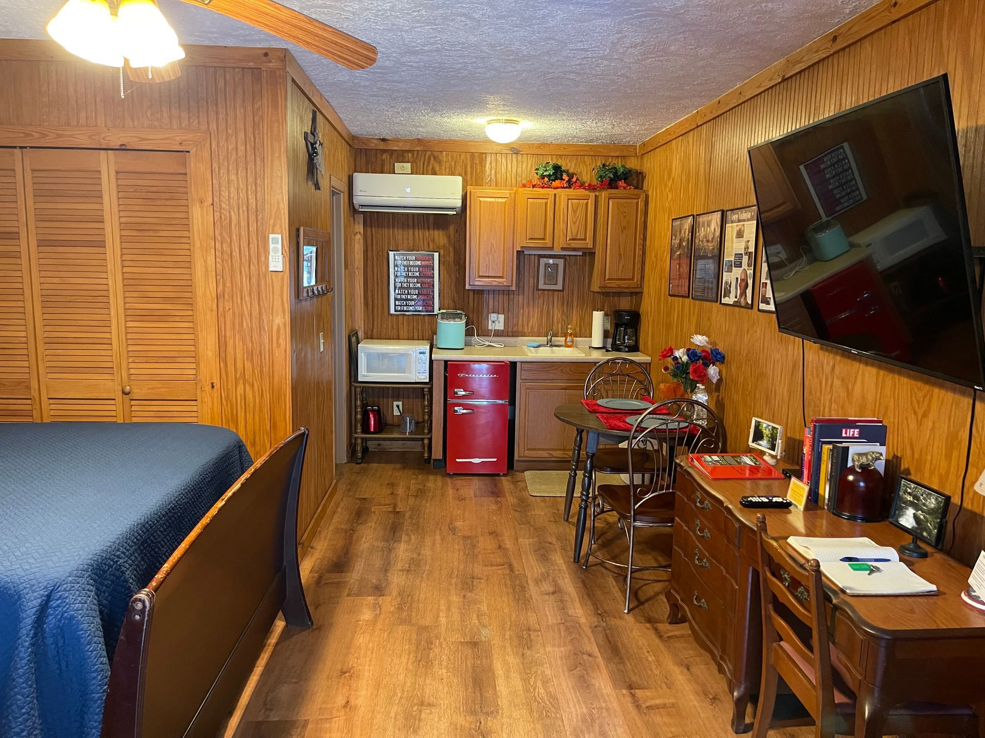 Cozy studio apartment with wood paneling, small kitchen, and bed; a flat-screen TV is mounted on the wall.
