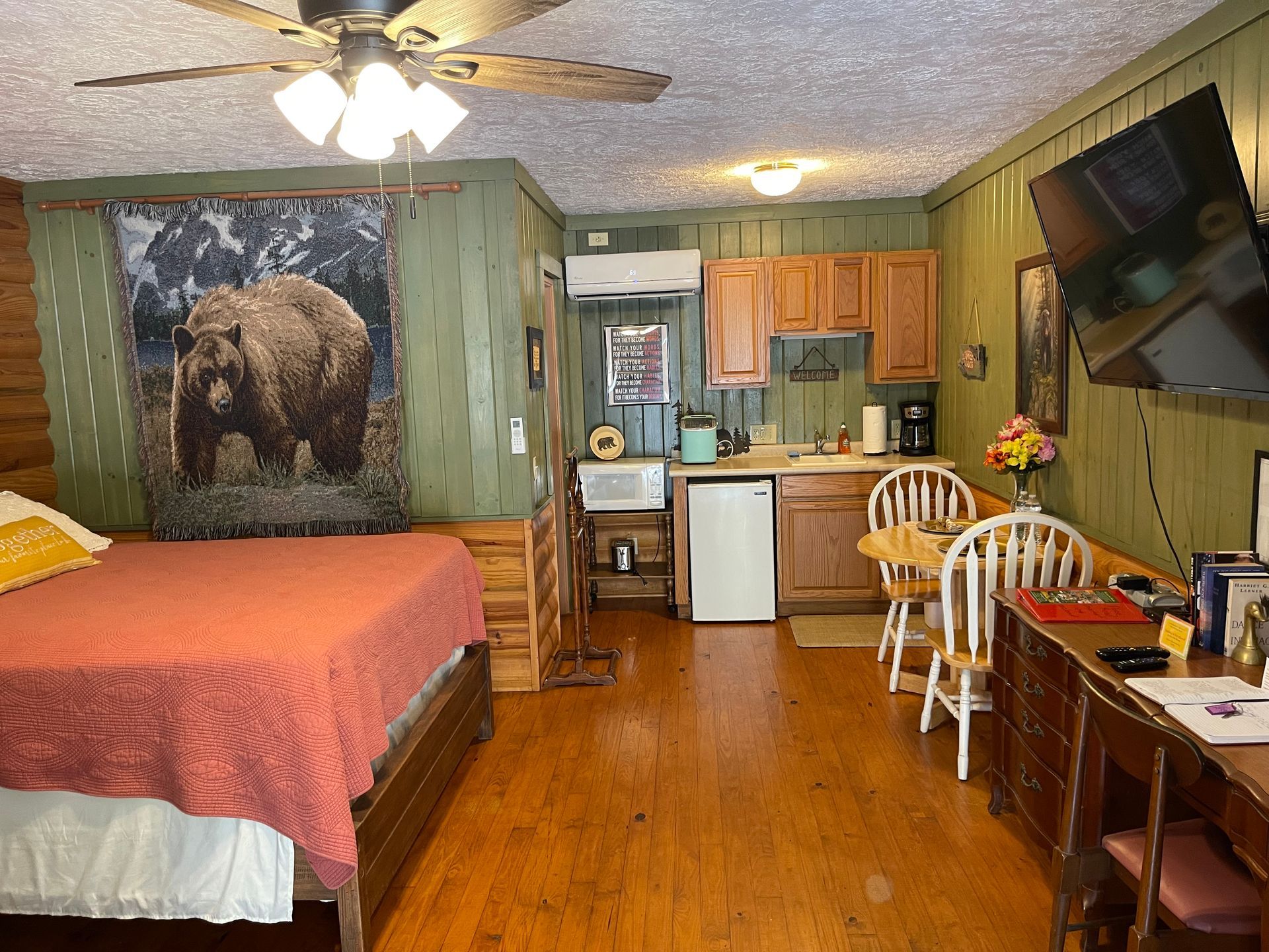 Cozy cabin room with a bed, kitchenette, bear tapestry, wooden table, and TV.
