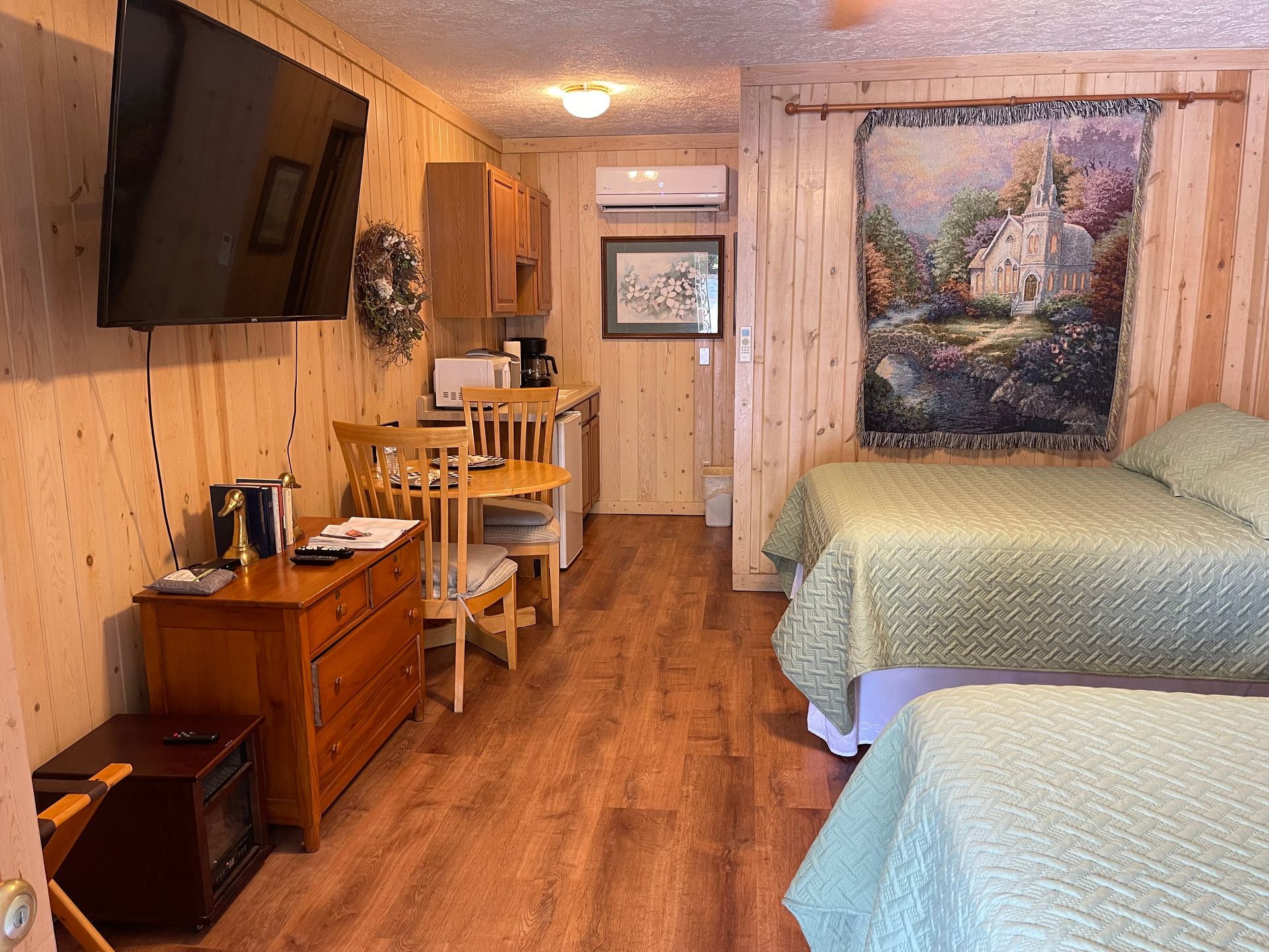 Small cabin interior with two beds, kitchenette, dresser, TV, and wall tapestry.