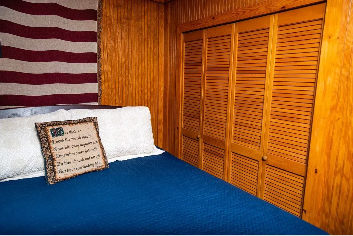 Bedroom with blue bed, wooden walls, and slatted closet doors. An American flag hangs above the bed.