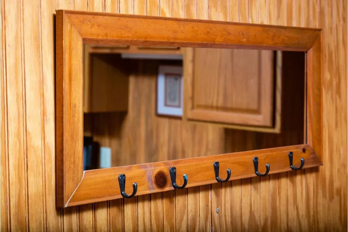 Mirror with hooks on a wood paneled wall; reflects a cabinet with an open door and a framed picture.