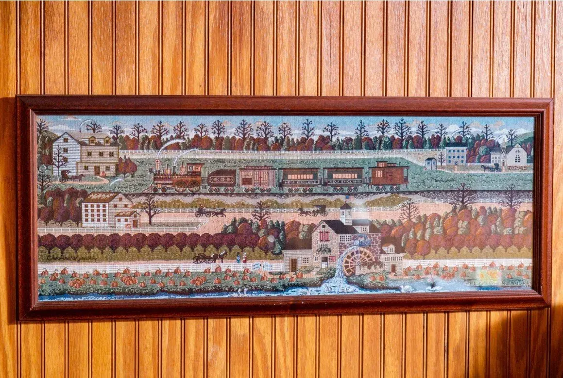 Framed tapestry depicts a train traveling through a landscape with buildings and trees, mounted on wood paneling.