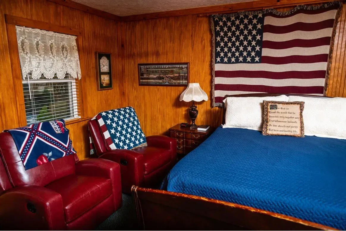 Bedroom with red leather chairs, American flag, Confederate flag, and wooden paneling.