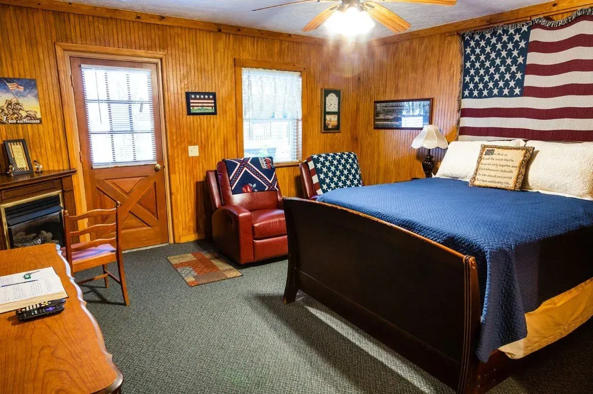 Cozy bedroom with wooden walls and an American flag. Features a bed, recliner, and desk.