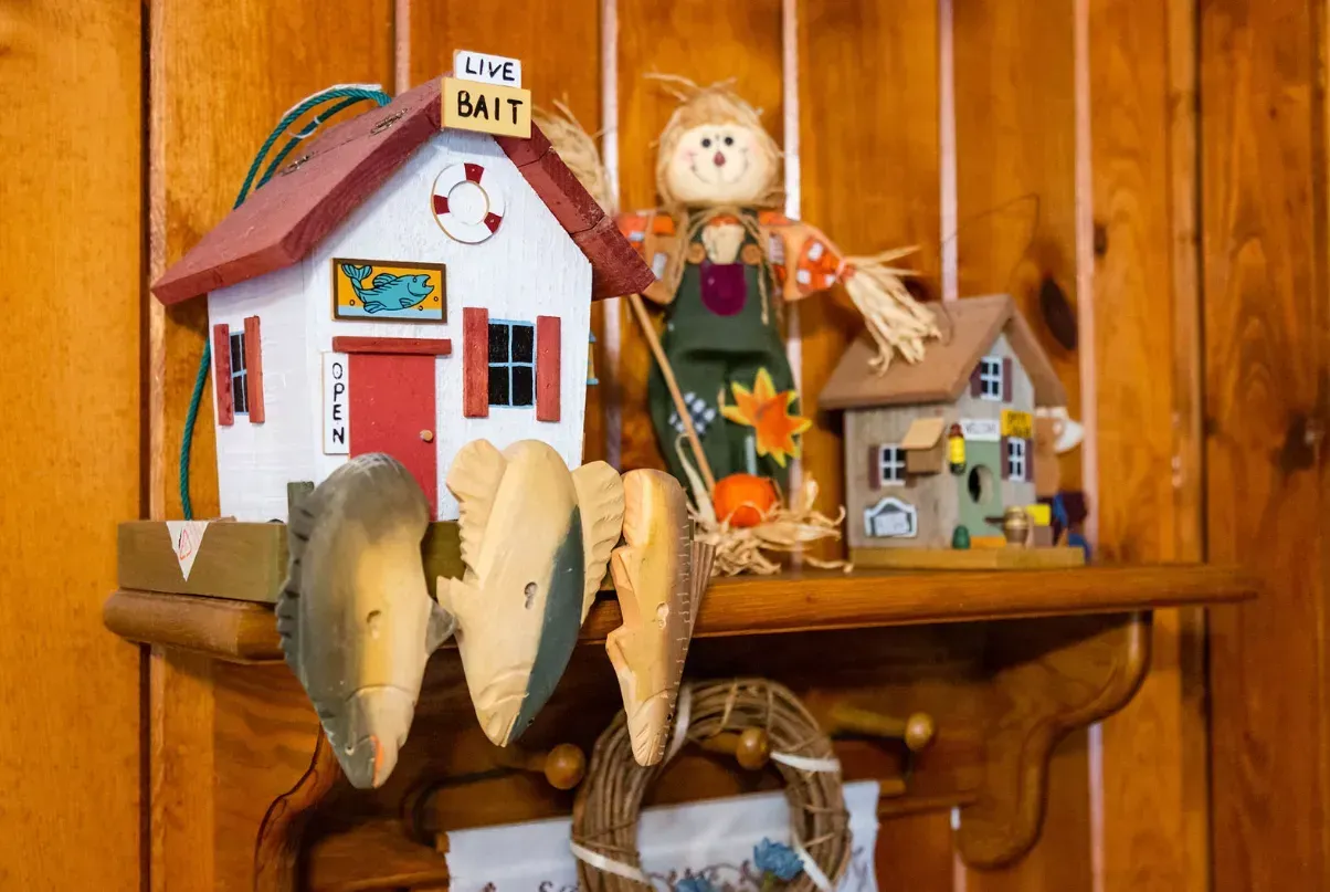Shelf with decorative houses, scarecrow, wooden fish, and wreath against wood paneling.