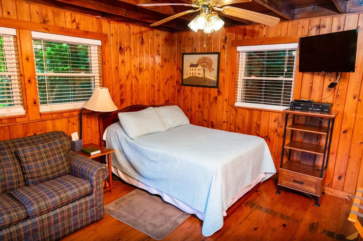 Cozy cabin bedroom with wood paneling, a bed, sofa, TV, and windows overlooking trees.