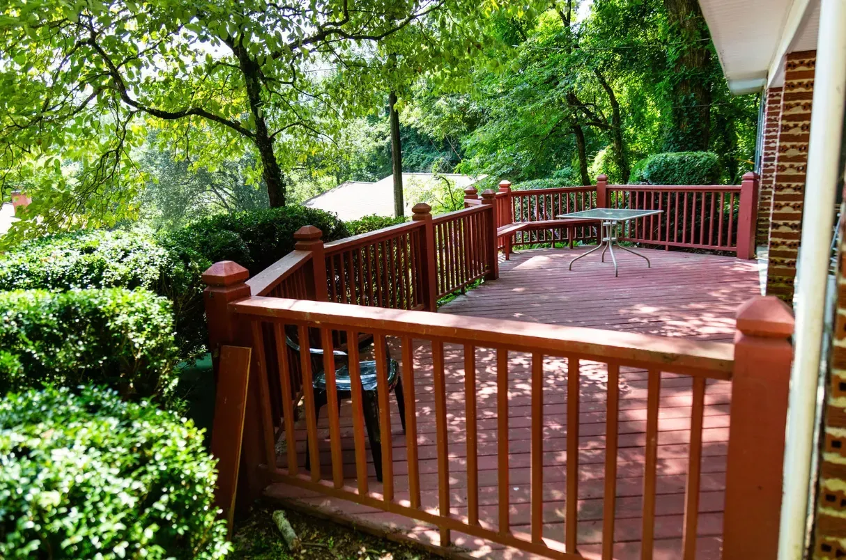 Red wooden deck with railings surrounded by greenery, table, and chairs.