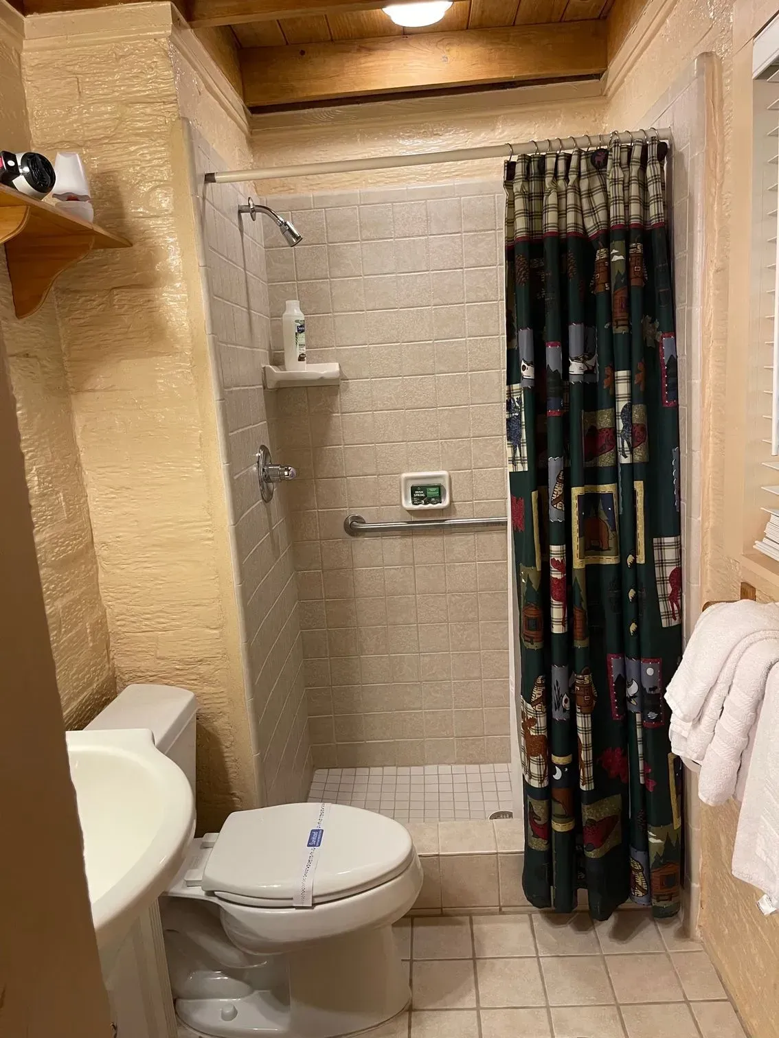 Bathroom with a toilet, shower, shower curtain, and sink. Beige walls, brown ceiling beams.